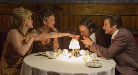 Rosalyn Rosenfeld (Jennifer Lawrence) insists on smelling her nail polish, Dolly Polito (Elisabeth Rohm), Irving Rosenfeld (Christian Bale) and Mayor Carmine Polito (Jeremy Renner) oblige inside Baron's Restauran. (Jennifer Lawrence jumpsuit, jewelry: vintage / Elisabeth Rohm dress: made for film, jewelry: vintage / Christian Bale suit: made for film, tie, shirt: vintage / Jeremy Renner suit, tie, shirt: vintage). Courtesy Columbia Pictures.