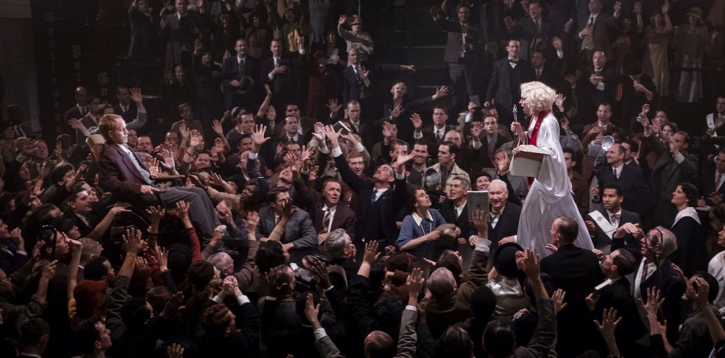 Sister Alice (Tatiana Maslany) works a miracle inside the Radiant Assembly of God interior, at the historic Trinity Auditorium location in downtown L.A. The four day shoot in September 2019 utilized more than 400 background actors each day, including a mass of congregants. Background cast with special improvisational skills were placed close to Maslany, near the stage, to be visible to camera as they portrayed being “saved,” speaking in tongues and other activity. Courtesy HBO.