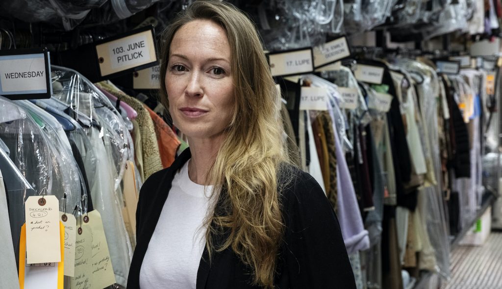 Costume Designer Emma Potter in front of one of countless racks of costumes on a location day. Note numbered tags on groups of clothing for particular repeat background cast, allowing quick access to the day’s specific wardrobe setup. Photo by Merrick Morton/HBO. 