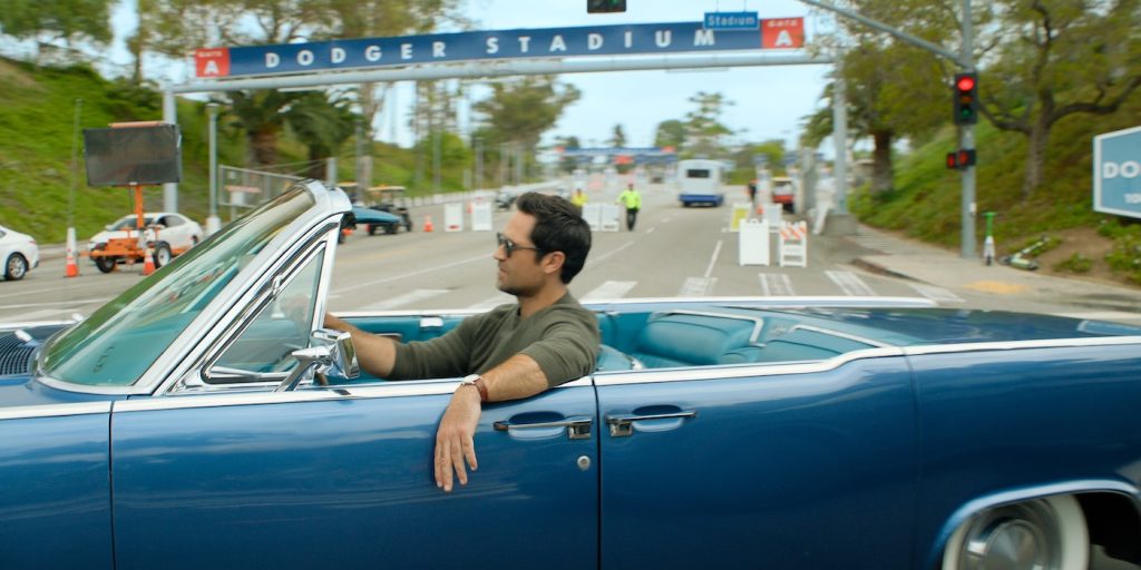 "Man driving a blue convertible near Dodger Stadium entrance on a sunny day, relaxed atmosphere, street scene with greenery, traffic lights, and stadium sign in the background."