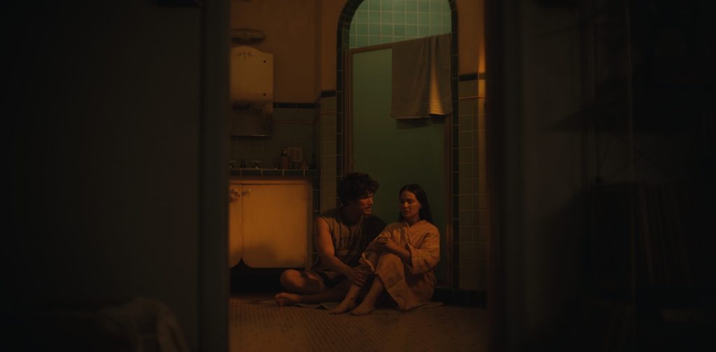 "Two people sit close together on a bathroom floor at night, lit by warm, soft lighting. The scene appears intimate and quiet, with vintage-style fixtures and a towel hanging over a glass door in the background."