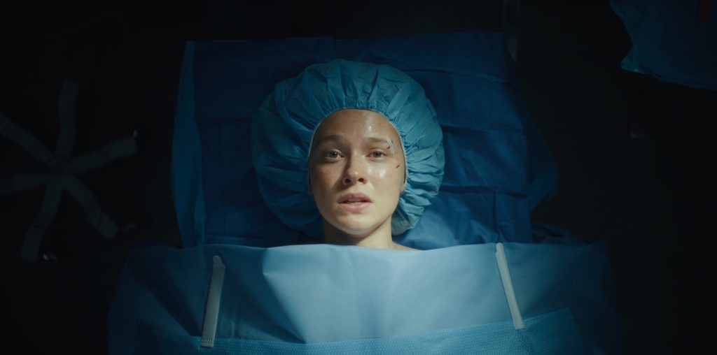 "Woman lying on an operating table under surgical drapes, wearing a blue surgical cap, looking up with a worried expression in a dimly lit hospital operating room."