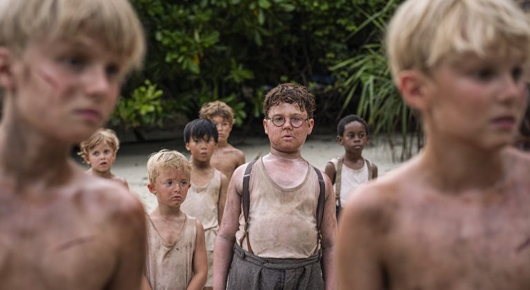 "A group of young boys with dirty faces and clothes stand on a sandy ground in a dense jungle, appearing tense and serious, likely stranded or lost in the wilderness."