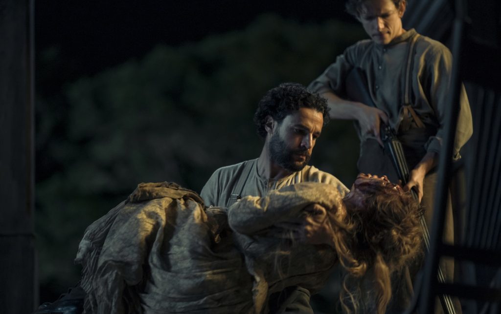 "A man holds an unconscious or injured woman in his arms at night, while another man with a rifle stands behind them outside, suggesting a dramatic or intense situation in a rural or historical setting."