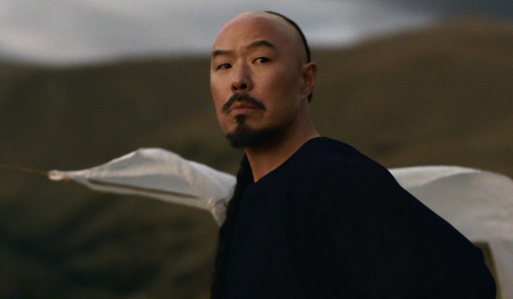 "A man with a mustache and goatee stands outdoors against rolling hills and cloudy sky, dressed in dark clothing with a white cloth behind him, looking toward the camera in a natural, rugged landscape."